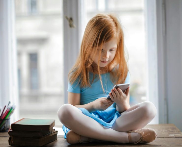 calm small ginger girl sitting on table and using smartphone in light living room