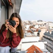 woman wearing maroon sweater standing on veranda using camera while smiling overlooking houses and buildings